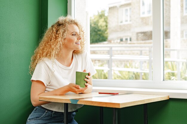 Кудрявая молодая женщина сидит за столом в кафе и расслабляется с чашкой кофе
