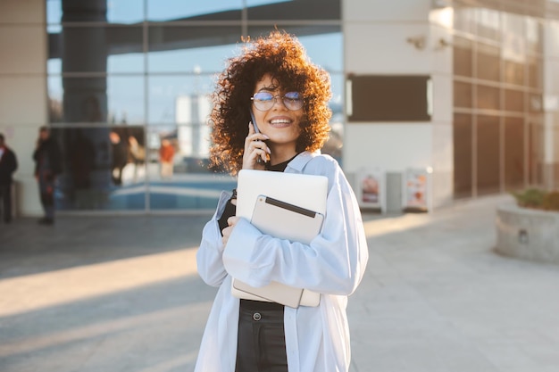 Curlyhaired business woman talking on mobile phone while holding a laptop and tablet in her han outs