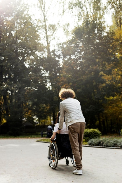 Curlyhaired 남자는 휠체어에 여자를 운반 다시보기 젊은 사람들이 거리를 걷고있다