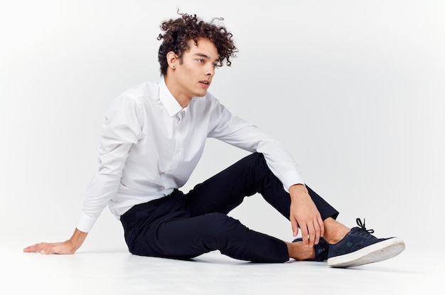 Curlyhaired man in shirt en broek en sneakers zit op de vloer binnenshuis zijaanzicht