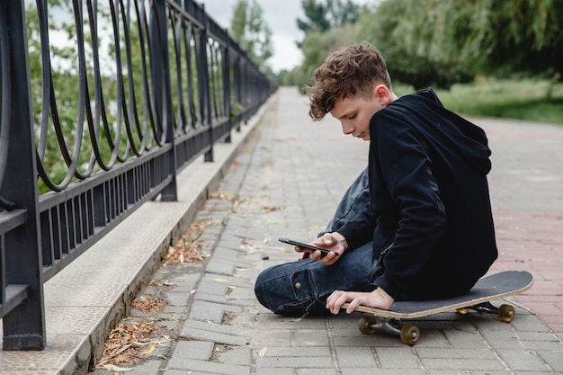 Un adolescente dai capelli ricci di aspetto europeo in felpa con cappuccio nera seduto su un vicolo su uno skateboard e ta...