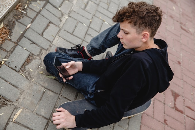 A curlyhaired teen of european appearance in black hoodie sitting on an alley on a skateboard and ta...