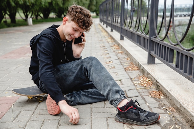 A curlyhaired teen of european appearance in black hoodie sitting on an alley on a skateboard and ta...