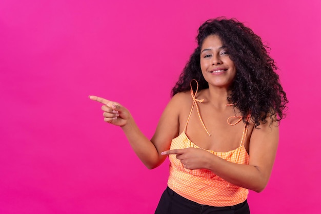 Donna dai capelli ricci in abiti estivi su uno sfondo rosa che punta a sinistra girato in studio