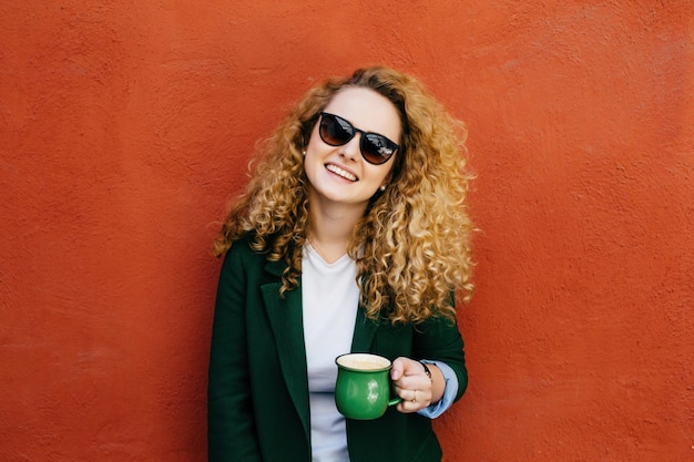 Curlyhaired woman sunglasses jacket cappuccino smile orange wall