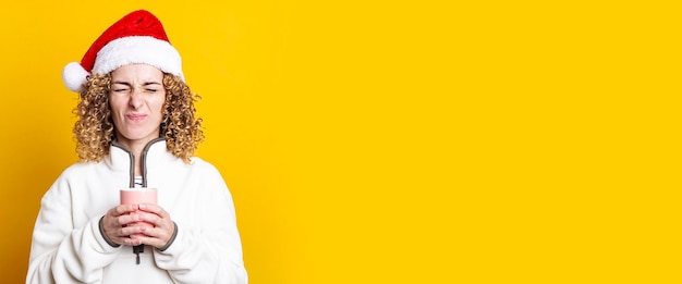 Curlyhaired young woman in santa claus hat with a cup on a yellow background Banner