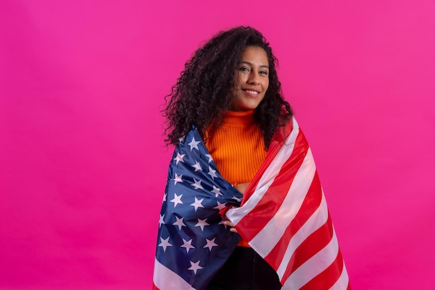 Curlyhairedvrouw die de vlag van de v.s. op een roze achtergrondstudioschot omhelzen