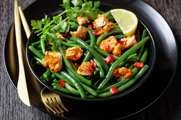 Curried zalm met sperziebonen en rode paprika geserveerd op een zwarte plaat met peterselie, citroenwig, op een donkere houten ondergrond met gouden bestek, bovenaanzicht, close-up