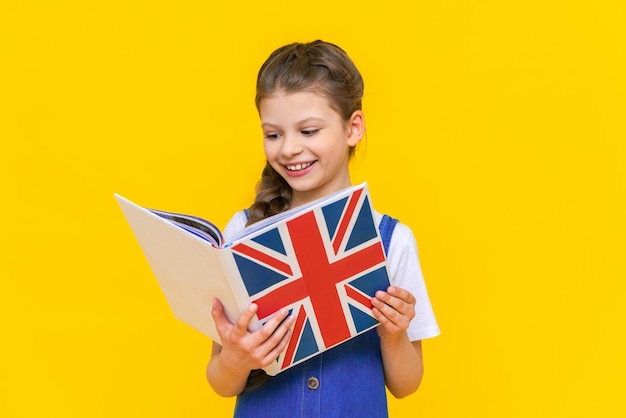 Cursussen Engels voor kinderen Een klein meisje houdt een boek vast met de vlag van Engeland en glimlacht breed Kinderonderwijs Talen leren Gele geïsoleerde achtergrond