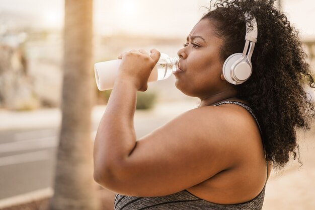 Curvy black woman drinking water after jog routine outdoor at city park - Focus on face