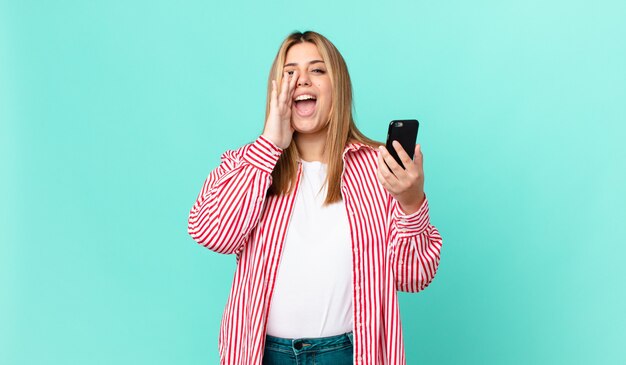 Curvy pretty blonde woman feeling happy,giving a big shout out with hands next to mouth and holding a smart phone