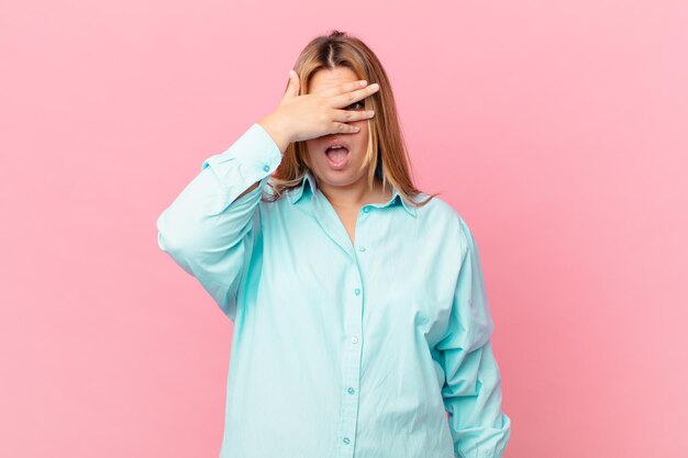 Curvy pretty blonde woman looking shocked, scared or terrified, covering face with hand