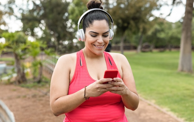 都市公園で屋外のジョギングルーチンをしながら携帯電話を使用してプレイリストの音楽を聞いている曲線美の女性-顔に焦点を当てる
