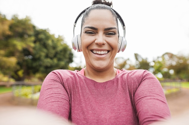 Curvy woman taking selfie with smartphone while doing jogging routine outdoor at city park - Plus size and workout exercises concept