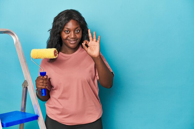 Curvy woman with paint roller and ladder cheerful and confident showing ok gesture