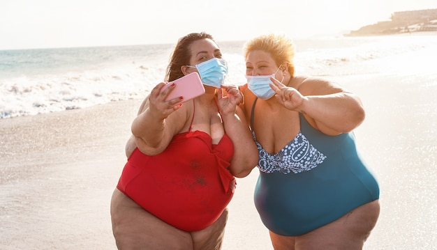 Curvy women friends taking selfie on the beach while wearing face mask for coronavirus spread prevention - Summer and healthcare concept