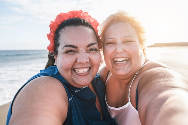 Amici di donne tutte curve che prendono selfie sulla spiaggia con il tramonto sullo sfondo - felice plus size corpo femminile divertendosi insieme - curve e concetto fiducioso