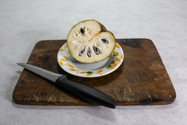 Custardappel typisch peruaans fruit de helft van het fruit in een porseleinen bord naast een keukenmes op een bruin houten bord en een witte achtergrond