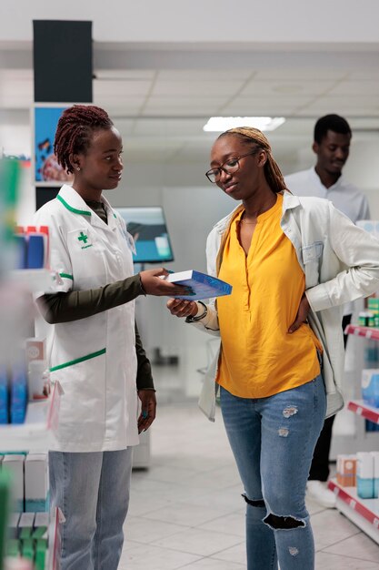 Customer buying back pain medication in drugstore, pharmacy\
consultant and buyer talking. african american woman client with\
backache standing, shopping, chatting with pharmacist, all black\
team