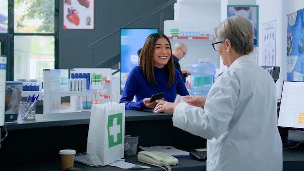 Customer at pharmacy counter, showing druggist prescribed cardiovascular pills picture on smartphone, ready to buy it. Elderly healthcare specialist selling client required medicinal product