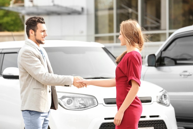 Customer and salesman shaking hands near new car outdoors