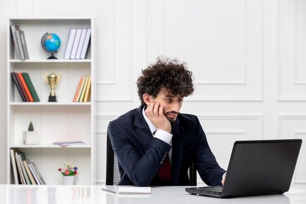 Customer service curly brunette young man in office suit and red tie with laptop looking at screen