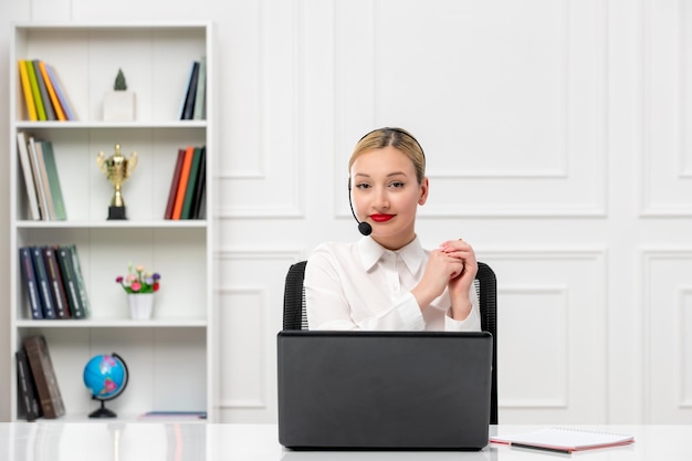 Customer service cute blonde girl in office shirt with headset\
and computer smiling and greeting