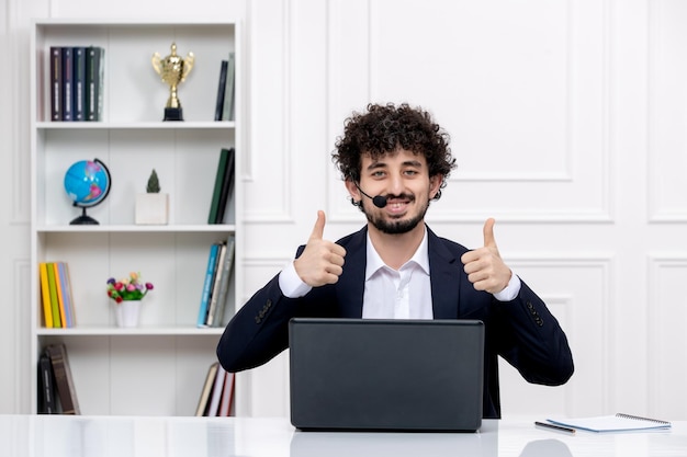 컴퓨터와 헤드셋이 있는 사무실 정장을 입은 귀여운 갈색 머리 남자가 행복하게 확인을 보여주고 있습니다.
