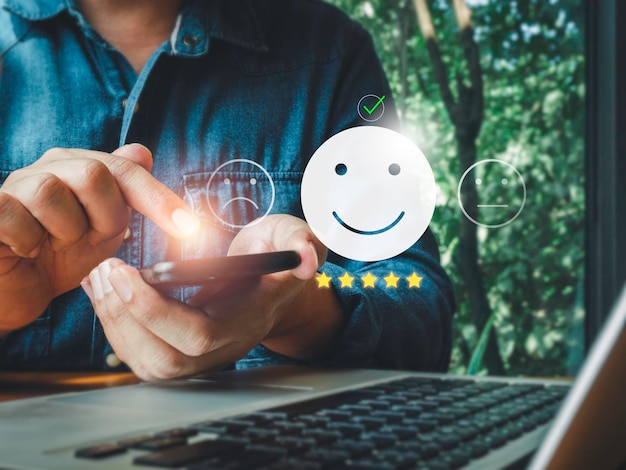 Photo customer service evaluation, feedback, client experience, and satisfaction survey concepts. checkmark on smiling happy face icon with five stars, rating by a man while using on phone and computer.