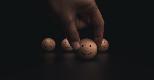Фото Оценка обслуживания клиентов, рейтинг, отзывы и концепция исследования удовлетворенности. рука, держащая смайлик счастливой улыбкой и размытые грустные лица на деревянном шаре на темном фоне.