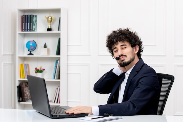 Customer service handsome curly man in office suit with computer and headset touching neck