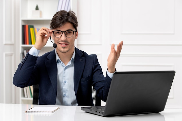 Bel ragazzo del servizio clienti con auricolare e laptop in giacca e cravatta che indossa gli occhiali e sorride