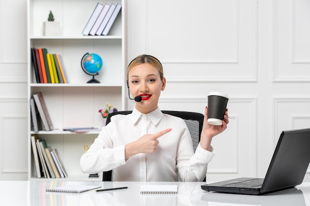 Foto servizio clienti bella ragazza bionda in camicia bianca con laptop e auricolare che tiene papercup nero