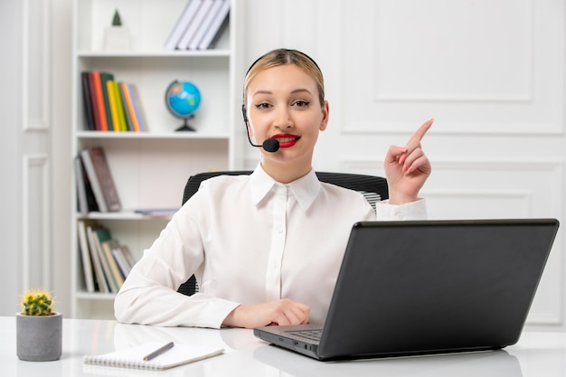 Customer service pretty blonde girl in white shirt with laptop and headset pointing up and smiling