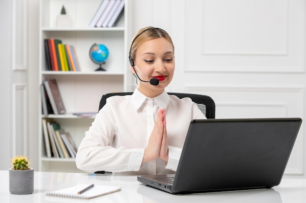 Customer service pretty blonde girl in white shirt with laptop and headset putting hands together