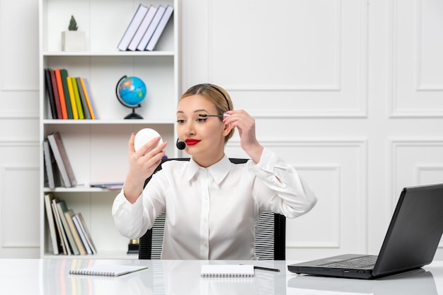 Customer service pretty blonde girl in white shirt with laptop and headset putting mascara on