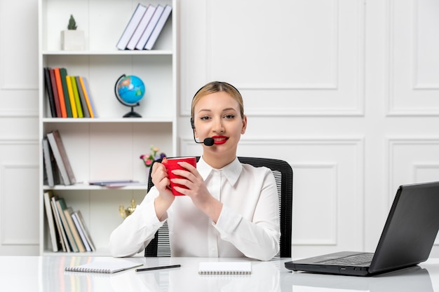 Photo customer service pretty blonde girl in white shirt with laptop and headset smiling with red cup