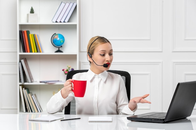 Customer service pretty blonde girl in white shirt with laptop and headset talking and holding cup