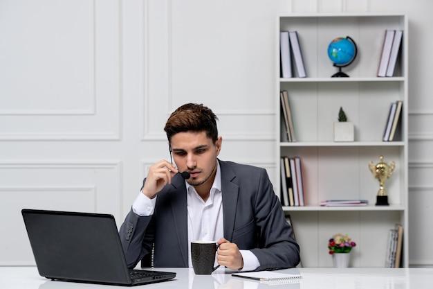 Customer service pretty gentleman with computer in grey office suit on the headset with coffee