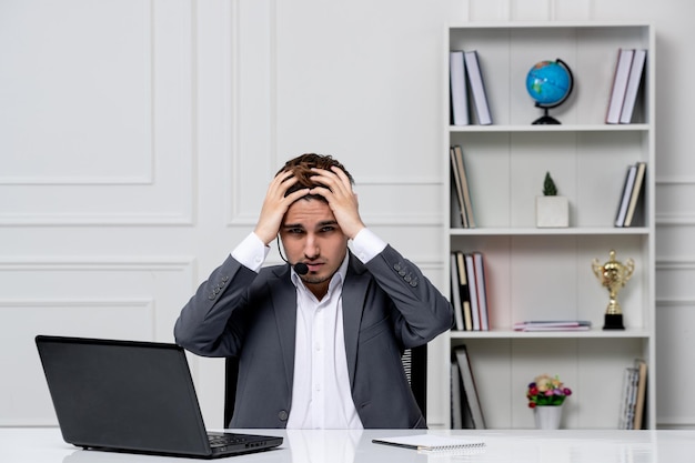 Customer service young cute guy in grey office suit with computer holding head extremely tired