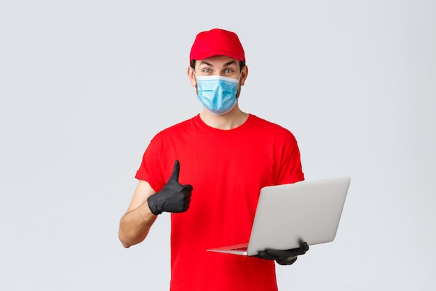 Customer support, covid-19 delivery packages, online orders processing concept. Enthusiastic courier in red uniform thumbs-up reading promo on webpage. Delivery guy hold laptop, wear mask and gloves