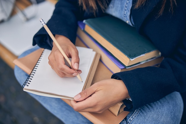 Cut off photo of female in jeans sitting and writing with pencil in notebook