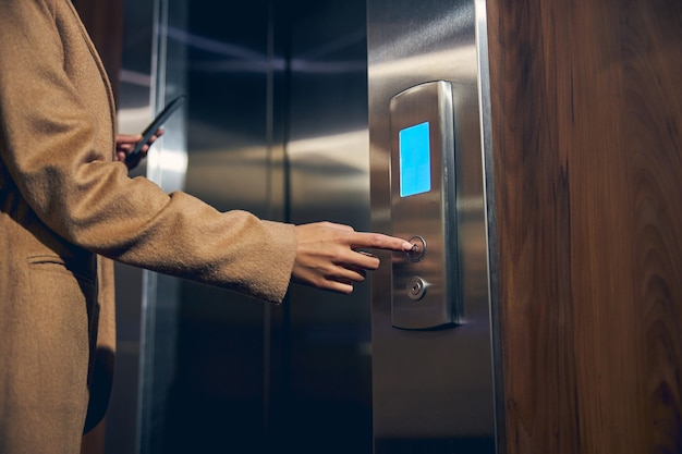 Cut off photo of lady in trendy coat pushing the button in the lift while holding her mobile