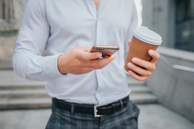 Cut view of body of young businessman stands and holds cup of coffee and phone.