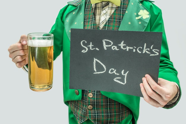 Cut view of man in green suit holding mug of beer and dark tablet with written words St. Patrick's Day. Isolated on grey .