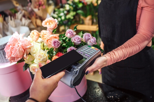 Cut view of man holding phone above money therminal. Lots of flowers and plants are behind.