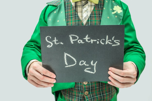 Отрежьте взгляд человека в зеленом костюме держа темную таблетку с днем st. patrick письменных слов. изолированные на сером.