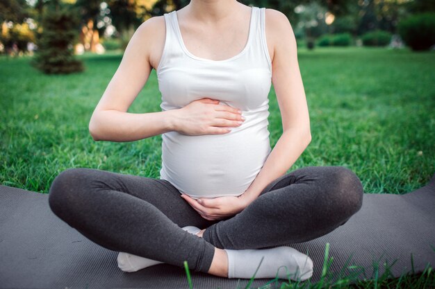 写真 公園でヨガの仲間に座っている若い妊婦のビューをカットします。彼女は腹の周りで手をつないでいます。モデルは蓮のポーズで座っています。