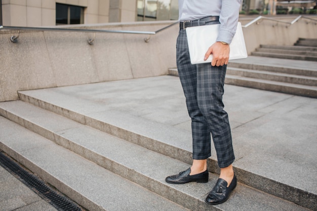 Cut view of stylish man stand and pose. He holds laptop with hand. Young man stands on steps.