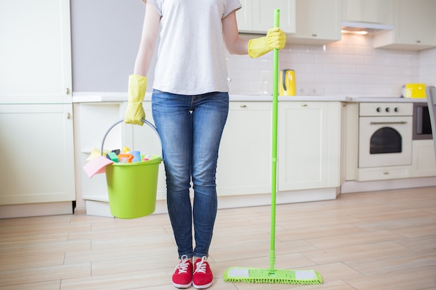 Cut view of woman stands in kitchen and holds stick with mop in one hand and bucket with cleaning equipment in the other one. Girl wears yellow gloves.